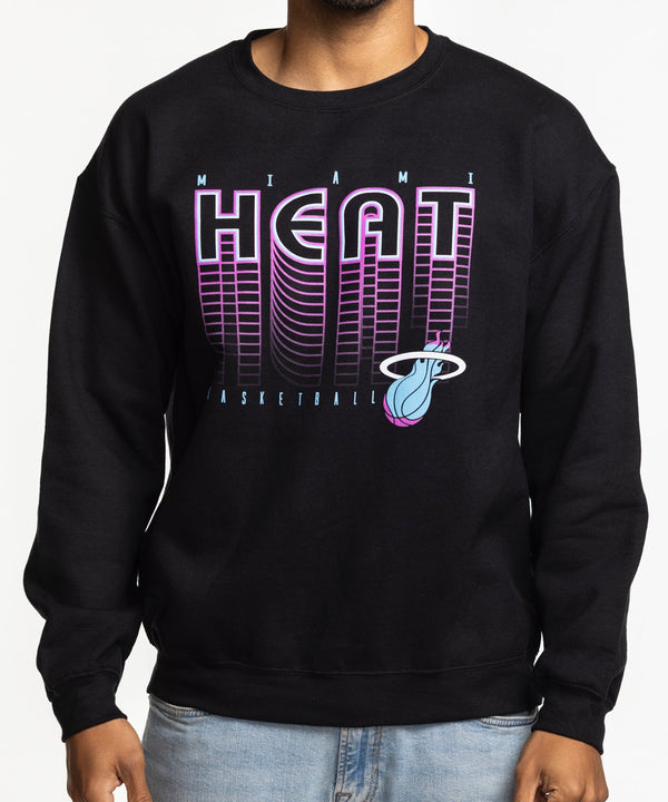 A man wearing the Item Of The Game Miami HEAT Original Vice Nights Retro Crewneck with a cotton-polyester blend and light blue jeans stands against a plain white background, smiling with one hand in his pocket.