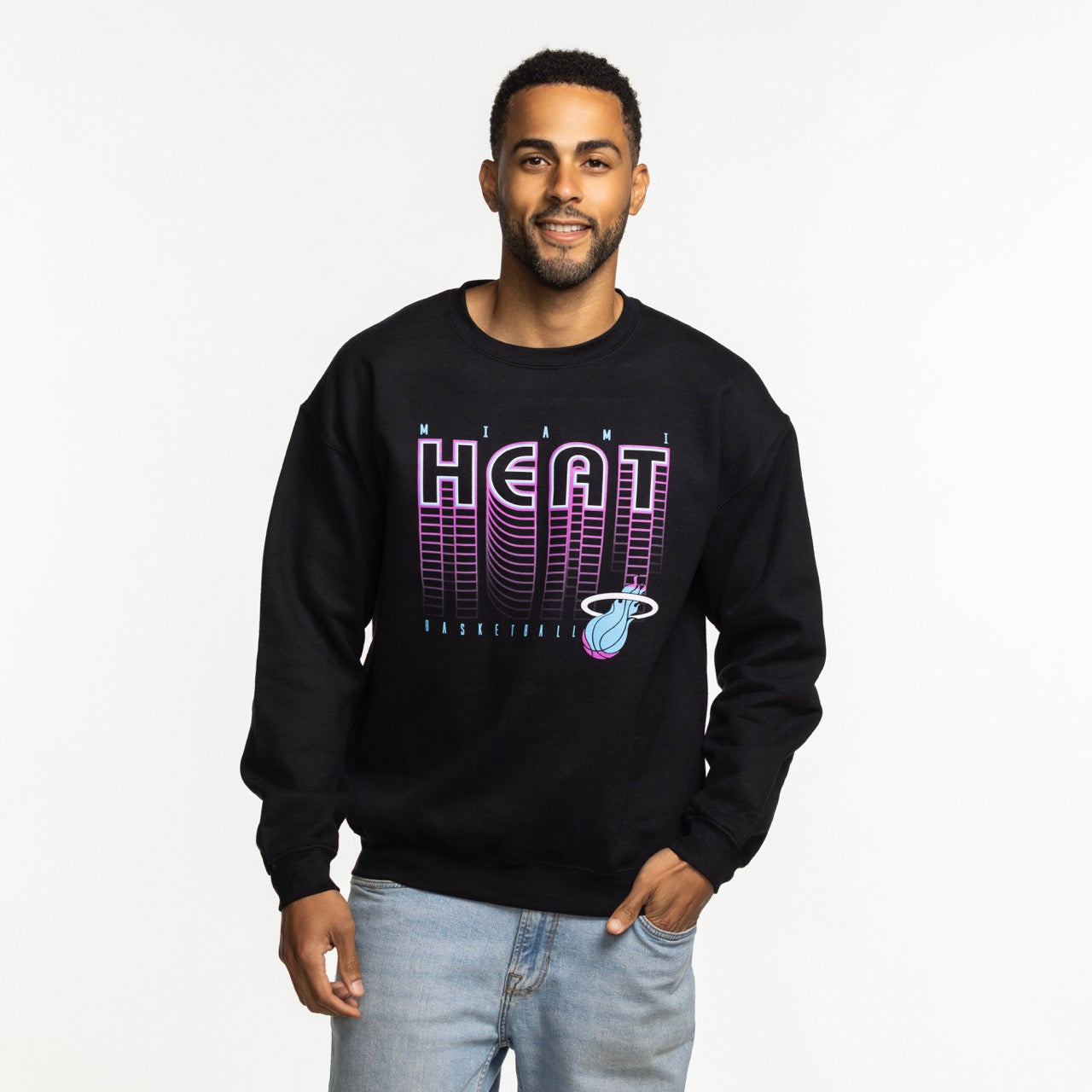 A man wearing the Item Of The Game Miami HEAT Original Vice Nights Retro Crewneck with a cotton-polyester blend and light blue jeans stands against a plain white background, smiling with one hand in his pocket.