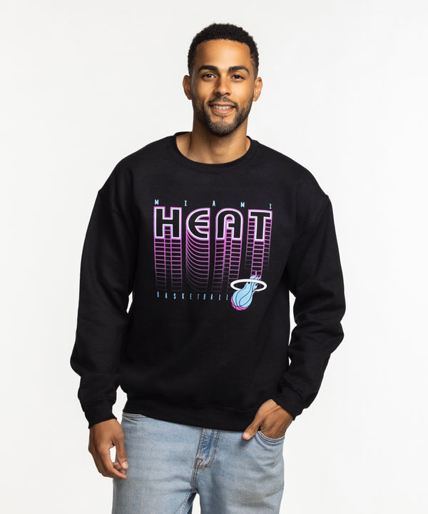 A man wearing the Item Of The Game Miami HEAT Original Vice Nights Retro Crewneck with a cotton-polyester blend and light blue jeans stands against a plain white background, smiling with one hand in his pocket.
