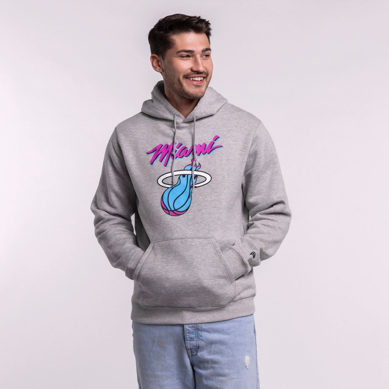 A smiling man with short brown hair models the New Era Miami HEAT Original Vice Grey Hoodie, featuring a colorful Miami flaming basketball graphic. He pairs it with light blue jeans and stands against a plain, light background. Cotton-polyester blend.