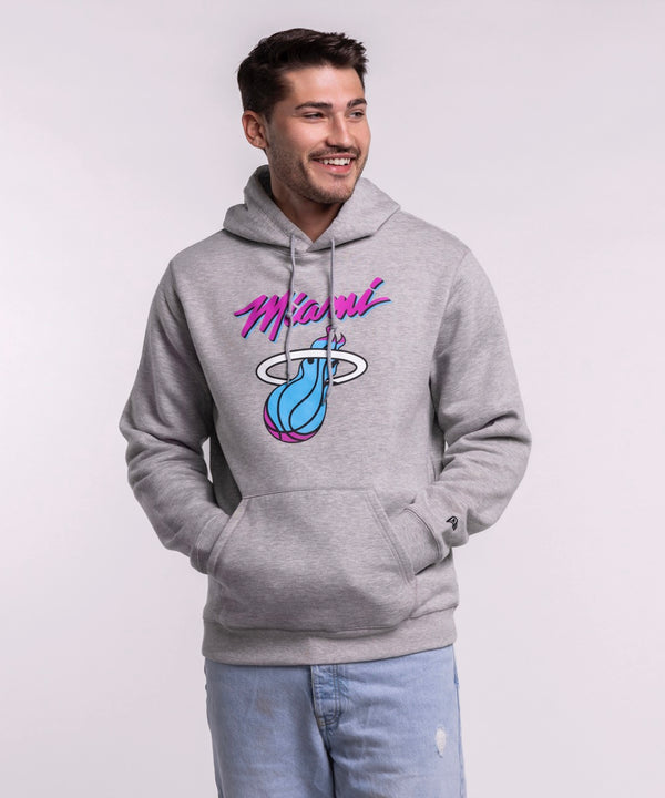 A smiling man with short brown hair models the New Era Miami HEAT Original Vice Grey Hoodie, featuring a colorful Miami flaming basketball graphic. He pairs it with light blue jeans and stands against a plain, light background. Cotton-polyester blend.