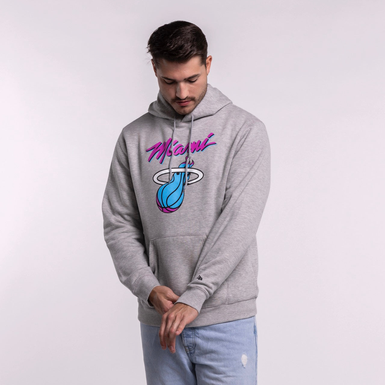 A man wearing the New Era Miami HEAT Original Vice Grey Hoodie by New Era, featuring a colorful team logo, and light blue ripped jeans stands against a plain background, looking down and adjusting his sleeve. Made from soft cotton-polyester.