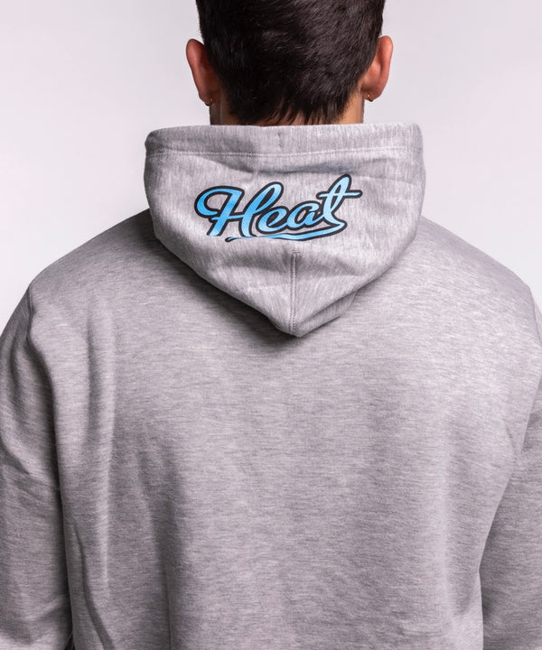 A smiling man with short brown hair models the New Era Miami HEAT Original Vice Grey Hoodie, featuring a colorful Miami flaming basketball graphic. He pairs it with light blue jeans and stands against a plain, light background. Cotton-polyester blend.
