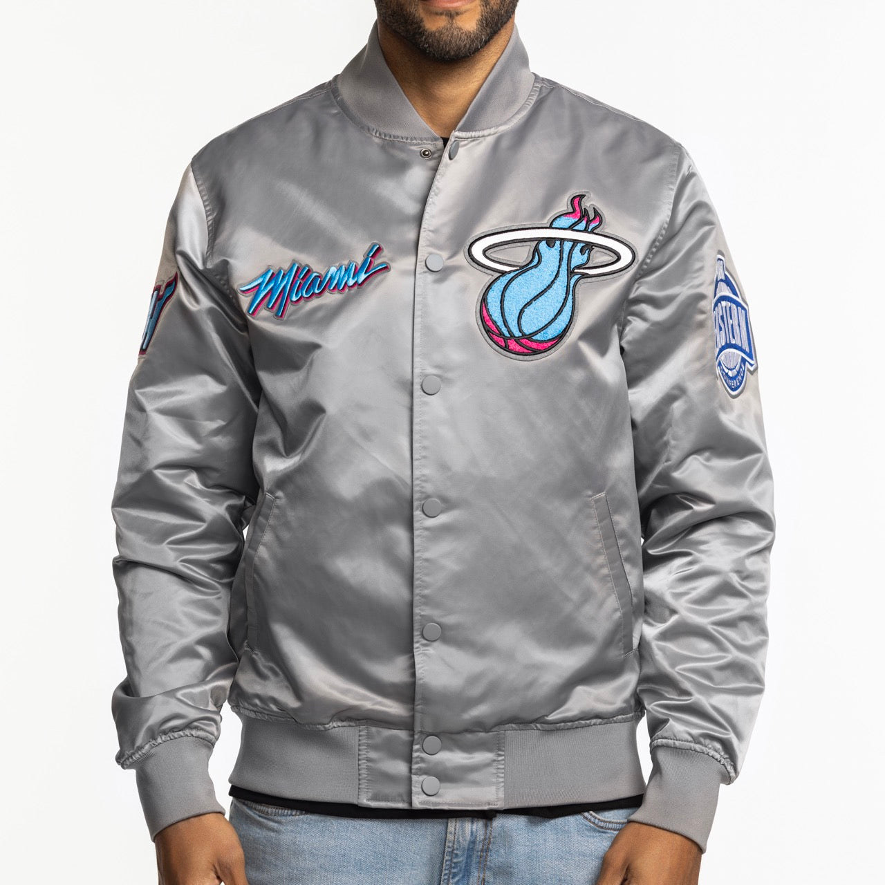 A person wearing the Pro Standard Miami HEAT Original Vice Nights Satin Jacket in shiny gray, standard fit, durable polyester with embroidered logos and text by Pro Standard, paired with light blue jeans on a plain white background. Machine washable.