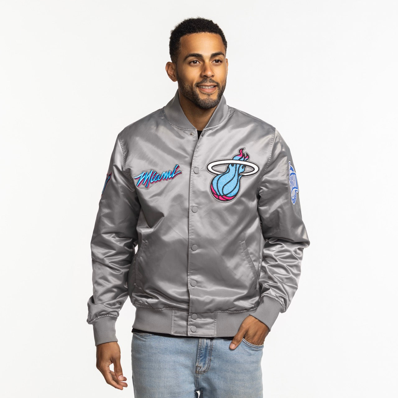 A man wearing a Pro Standard Miami HEAT Original Vice Nights Satin Jacket and light blue jeans stands smiling against a plain white background.