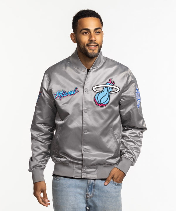 A man wearing a Pro Standard Miami HEAT Original Vice Nights Satin Jacket and light blue jeans stands smiling against a plain white background.