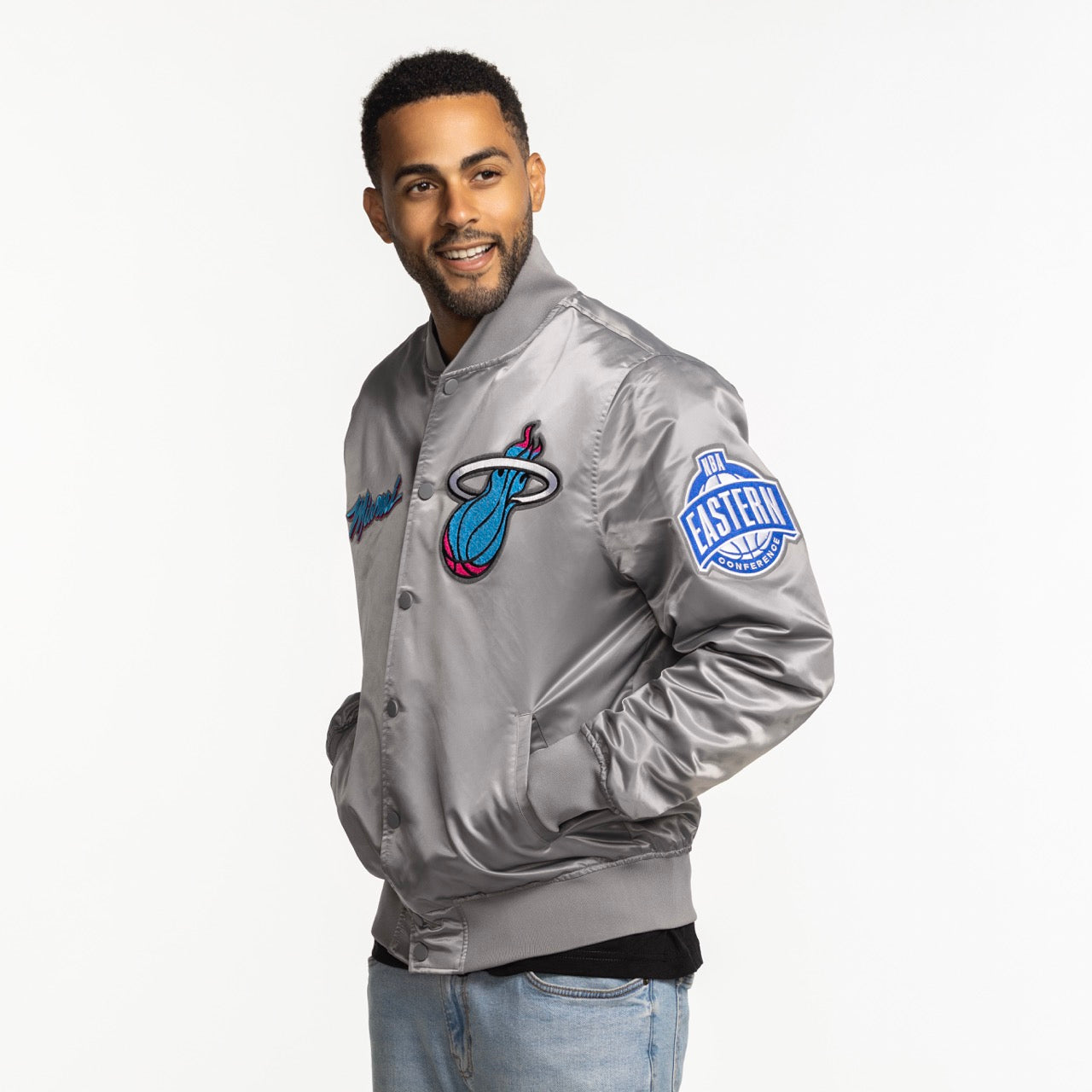 A man stands smiling with his hand in his pocket, wearing the Pro Standard Miami HEAT Original Vice Nights Satin Jacket—a shiny silver polyester bomber with a blue Eastern Conference patch on the sleeve. Standard fit; machine washable for easy care.