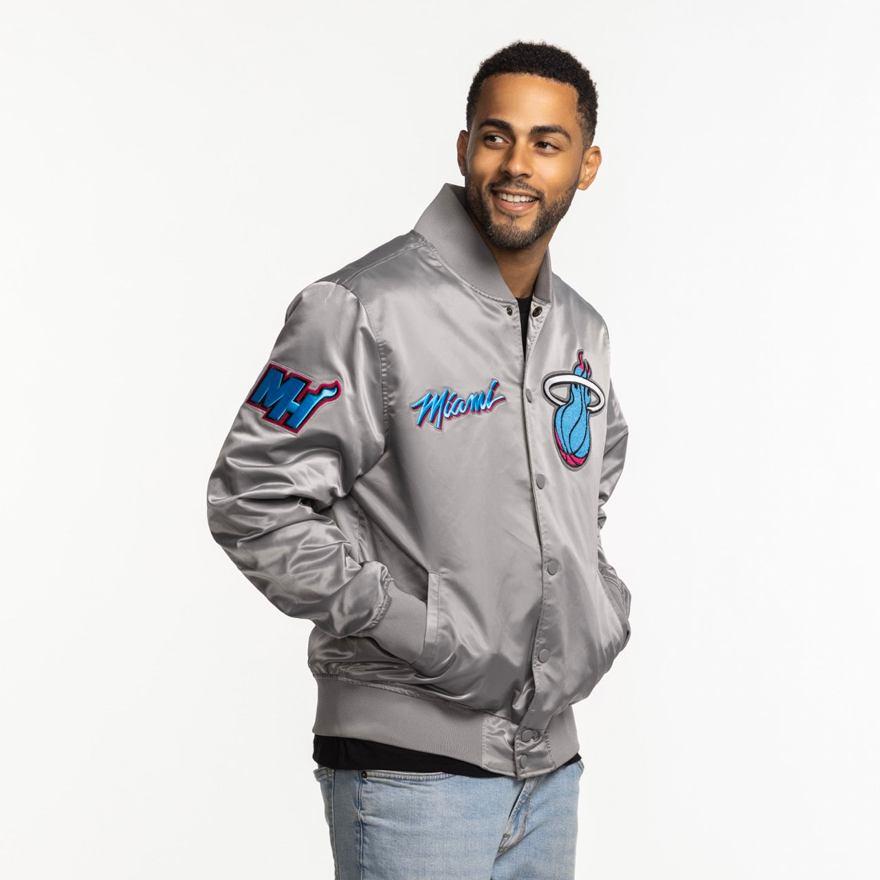 A man wearing the Pro Standard Miami HEAT Original Vice Nights Satin Jacket by Pro Standard, featuring colorful team logos and a shiny silver finish, pairs it with light blue jeans and smiles slightly with hands in pockets against a white background.