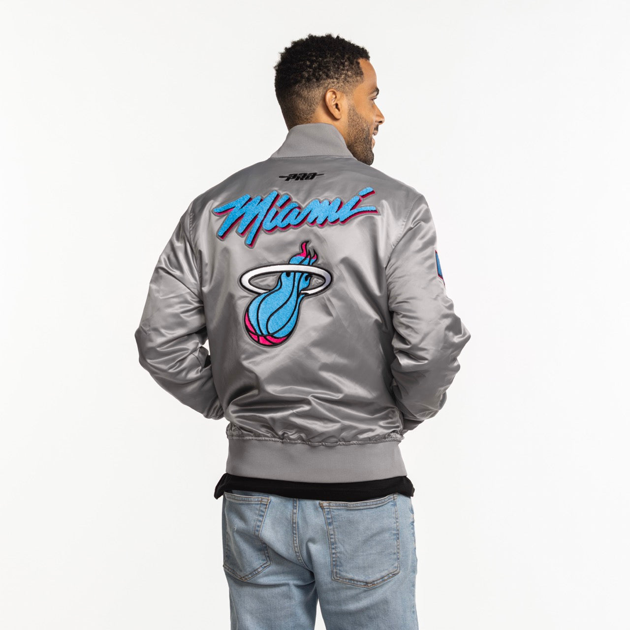 A man with short hair and blue jeans faces away, wearing a Pro Standard Miami HEAT Original Vice Nights Satin Jacket—an easy-care, machine-washable polyester bomber featuring colorful Miami text and the team's logo on the back.