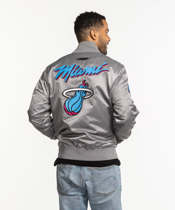 A man wearing a Pro Standard Miami HEAT Original Vice Nights Satin Jacket and light blue jeans stands smiling against a plain white background.