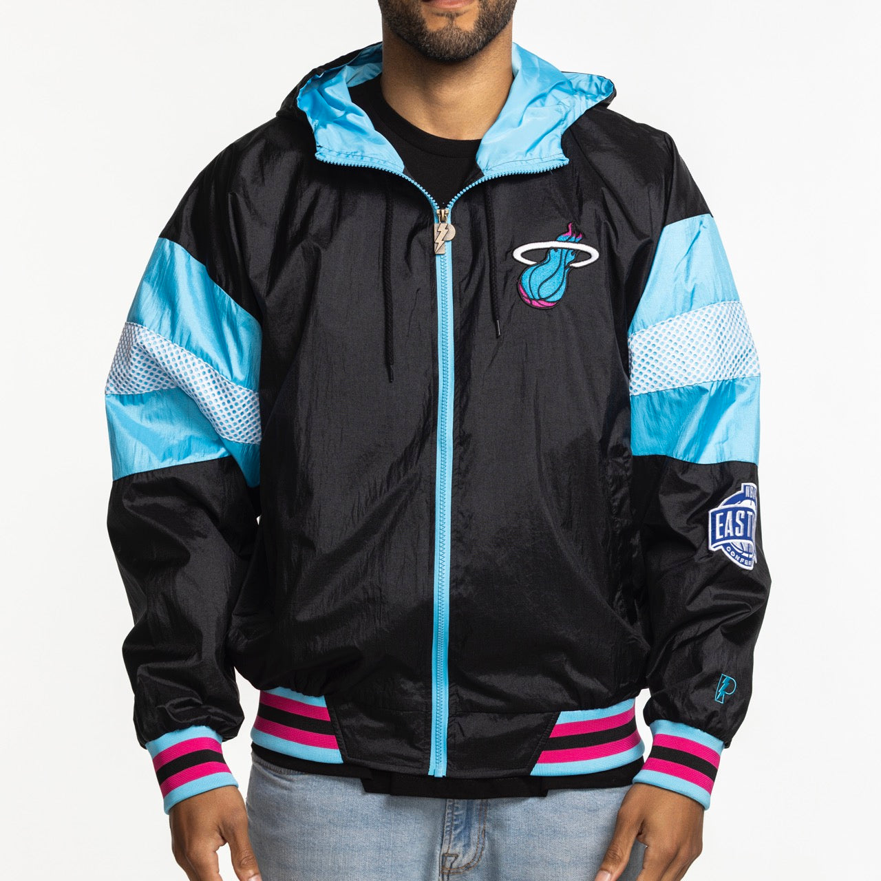 A person wearing a G-III Sports Miami HEAT Original Vice Nights Legacy Jacket with black, teal, pink, and white accents, NBA East patch on the sleeve, and light blue jeans. The face is not visible.