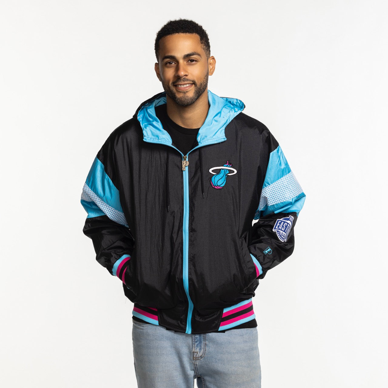 A man wears a G-III Sports Miami HEAT Original Vice Nights Legacy Jacket in black, pink, and blue with polyester lining, paired with light blue jeans, and smiles against a plain white background.
