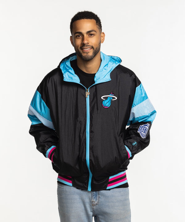 A man wears a G-III Sports Miami HEAT Original Vice Nights Legacy Jacket in black, pink, and blue with polyester lining, paired with light blue jeans, and smiles against a plain white background.