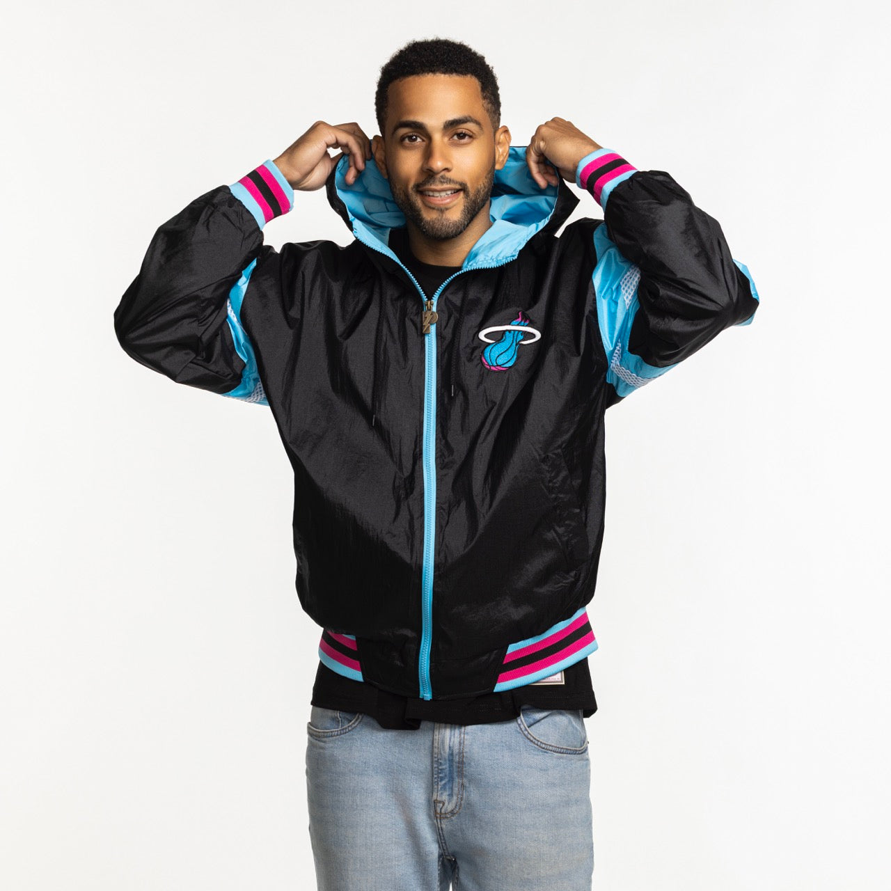 A man smiles, adjusting the hood of a G-III Sports Miami HEAT Original Vice Nights Legacy Jacket in black, pink, and blue with a polyester lining, paired with light blue jeans. The background is plain white.