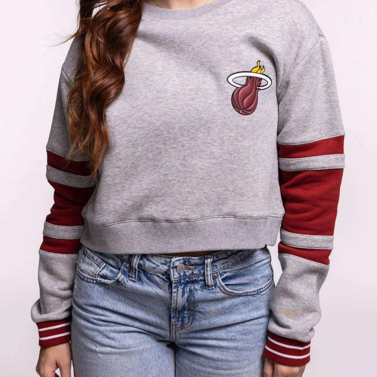 A person wears the Stadium Essentials Miami HEAT Women's Cropped Crewneck, a relaxed gray sweatshirt with a Miami Heat logo and red-striped sleeves, paired with light blue jeans. Made from a cotton-polyester blend. Face not shown. Brand: Stadium Essentials.