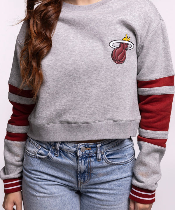 A young woman with long brown hair wears the Stadium Essentials Miami HEAT Women's Cropped Crewneck, a relaxed gray sweatshirt with red stripes, and blue jeans. Made by Stadium Essentials from soft cotton-polyester. Machine wash cold. She smiles slightly at the camera.