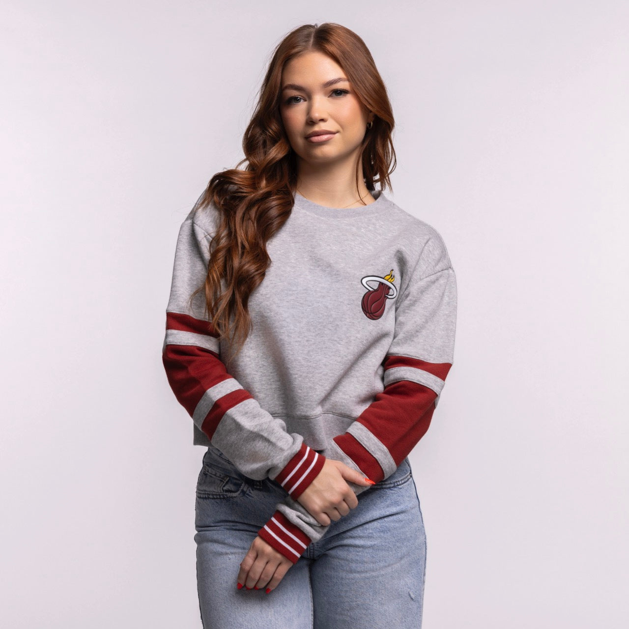 A young woman with long brown hair wears the Stadium Essentials Miami HEAT Women's Cropped Crewneck, a relaxed gray sweatshirt with red stripes, and blue jeans. Made by Stadium Essentials from soft cotton-polyester. Machine wash cold. She smiles slightly at the camera.