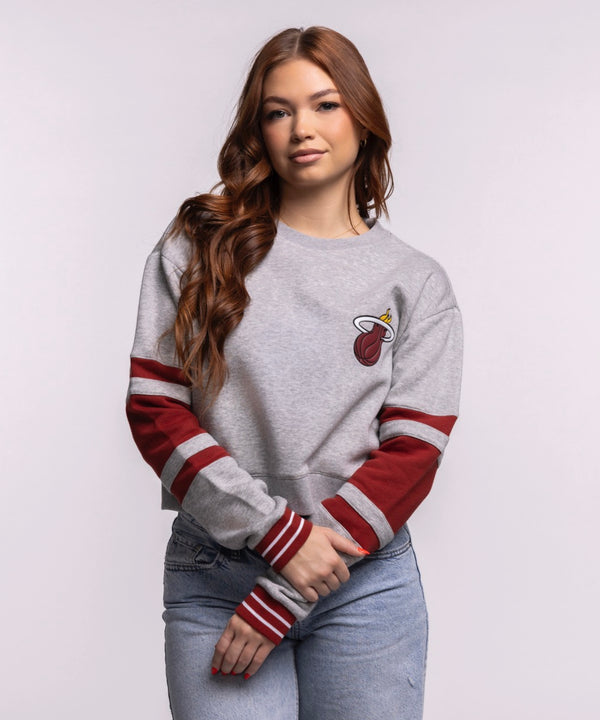 A young woman with long brown hair wears the Stadium Essentials Miami HEAT Women's Cropped Crewneck, a relaxed gray sweatshirt with red stripes, and blue jeans. Made by Stadium Essentials from soft cotton-polyester. Machine wash cold. She smiles slightly at the camera.