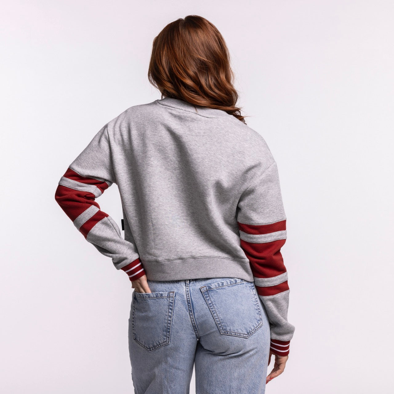 A woman with long, wavy brown hair faces away, wearing a Stadium Essentials Miami HEAT Women's Cropped Crewneck in light gray with maroon sleeve stripes and light blue high-waisted jeans, standing against a plain light gray background.