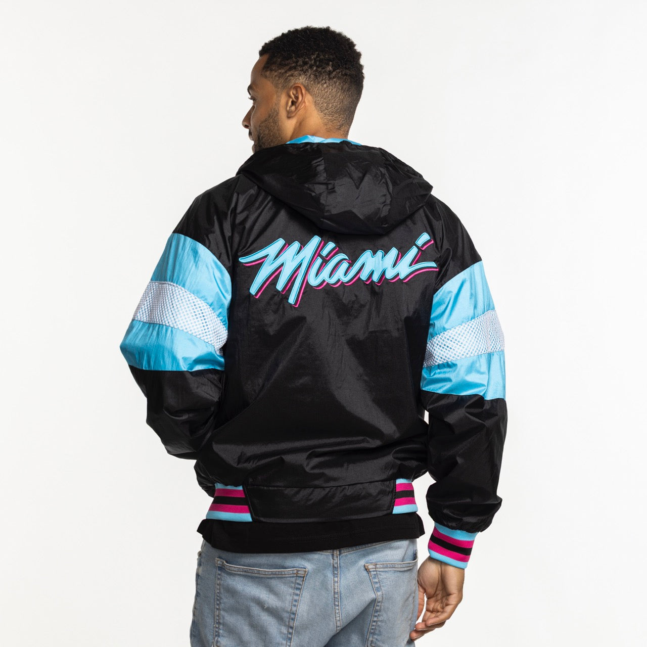 A man with short hair, seen from behind, wears the G-III Sports Miami HEAT Original Vice Nights Legacy Jacket—a black nylon hooded jacket with blue and pink accents, 