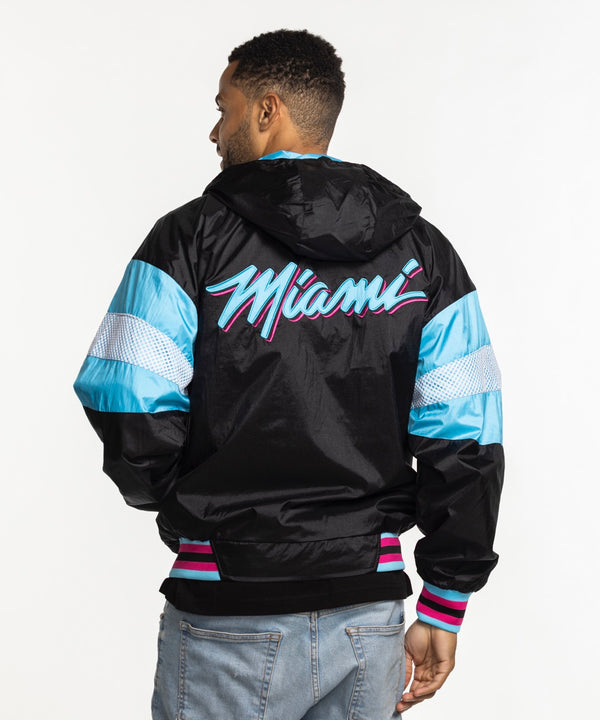 A man wears a G-III Sports Miami HEAT Original Vice Nights Legacy Jacket in black, pink, and blue with polyester lining, paired with light blue jeans, and smiles against a plain white background.