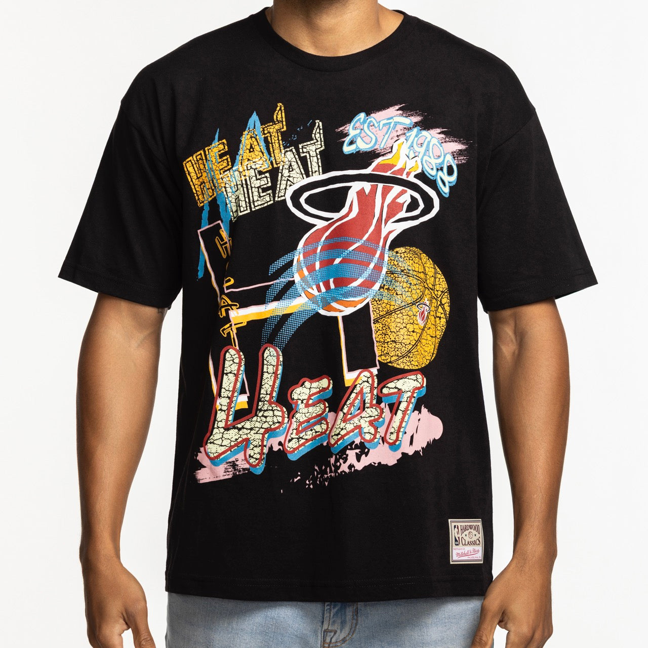 Someone wears the Mitchell & Ness Miami HEAT Mixed Up Tee, a black 100% cotton T-shirt by Mitchell & Ness featuring a colorful Miami Heat graphic, bold HEAT logo, EST. 1988, and abstract patterns. Machine wash cold for best results.