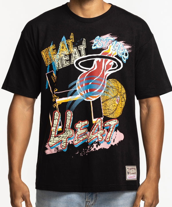 A man wears the Mitchell & Ness Miami HEAT Mixed Up Tee by Mitchell & Ness with light blue jeans, smiling against a plain white background.