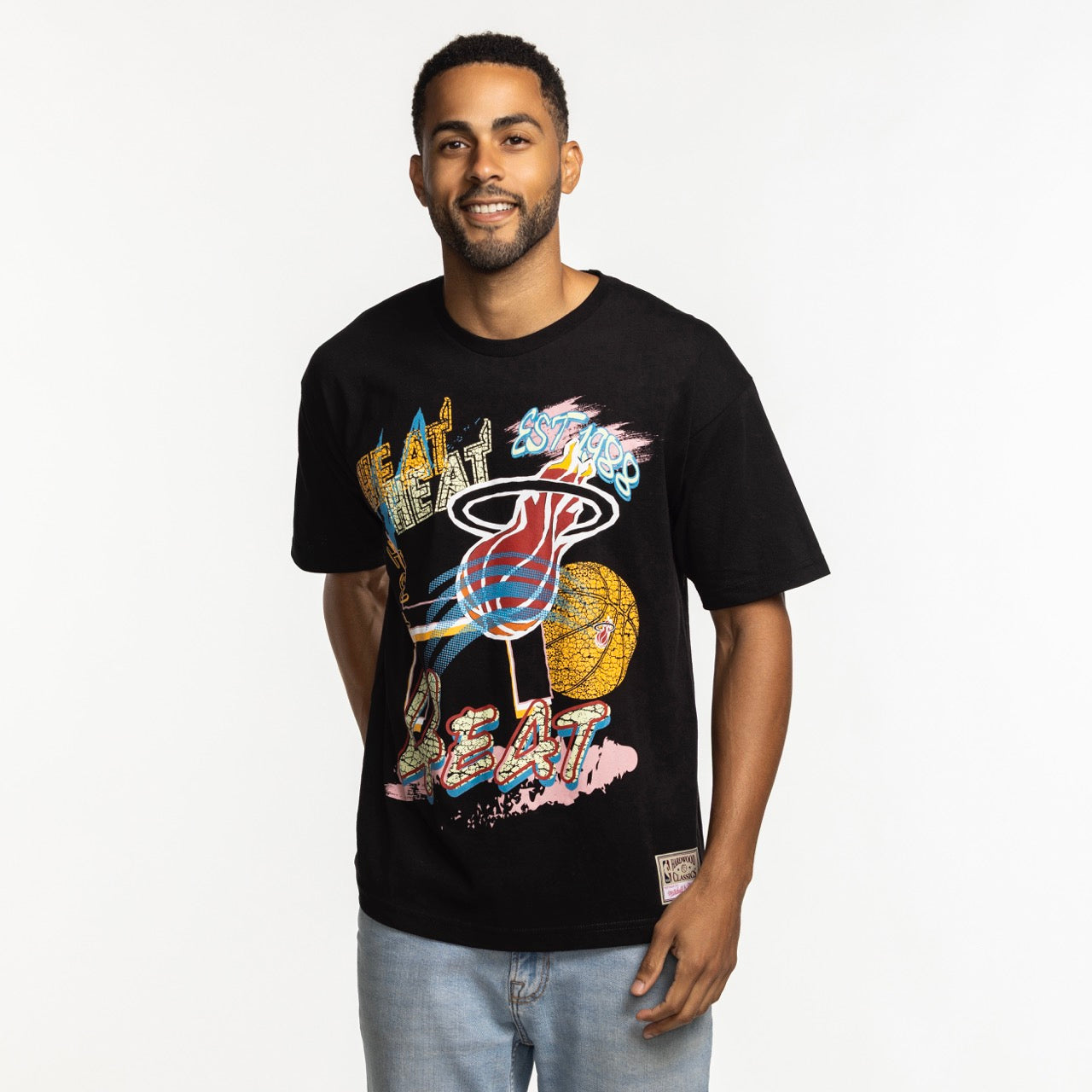 A man wears the Mitchell & Ness Miami HEAT Mixed Up Tee by Mitchell & Ness with light blue jeans, smiling against a plain white background.