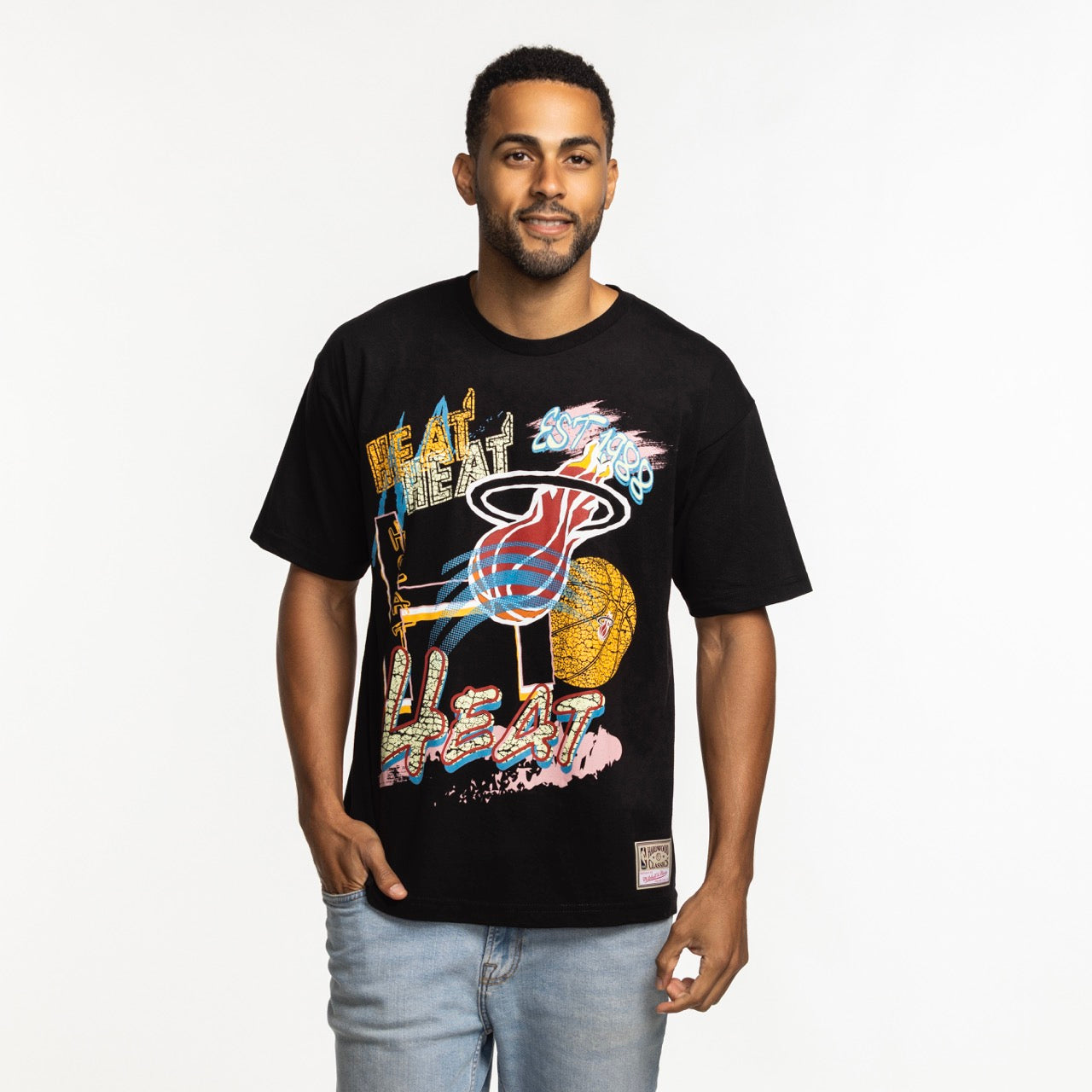 A man with short hair and a beard smiles with one hand in his pocket, wearing the Mitchell & Ness Miami HEAT Mixed Up Tee and light blue jeans, standing against a plain white background.