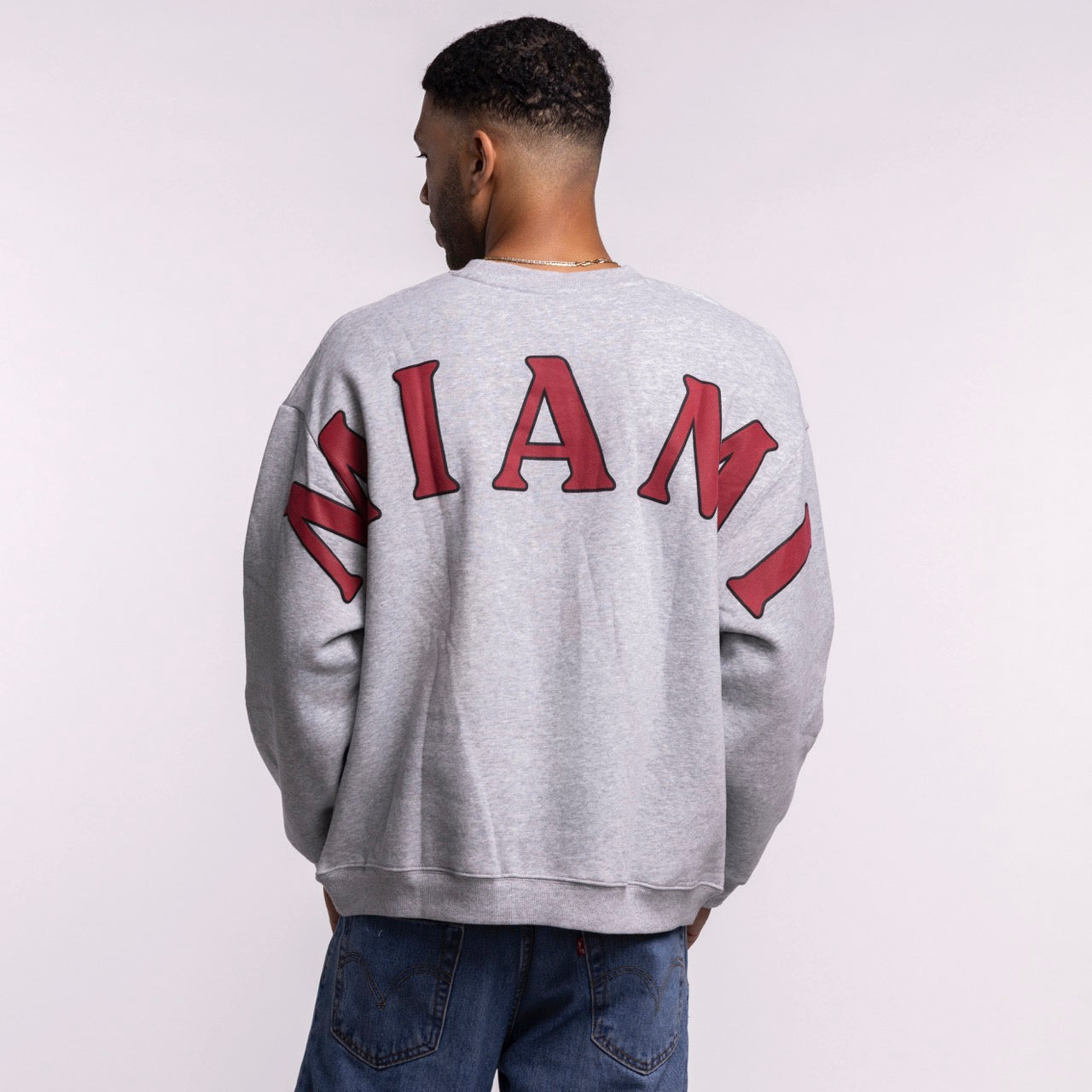 A person wearing the Hype and Vice Miami HEAT Oversized Crew—a gray, oversized sweatshirt by Hype and Vice with bold red MIAMI on the back—stands with their back to the camera, paired with blue jeans. Soft cotton-poly blend; hand wash cold recommended.