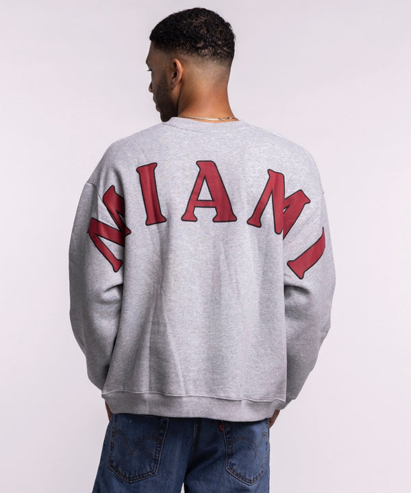 A woman with long brown hair wears the Hype and Vice Miami HEAT Oversized Crew by Hype and Vice, a gray cotton-polyester blend sweatshirt, paired with light blue jeans, standing against a plain background. Hand wash cold recommended.