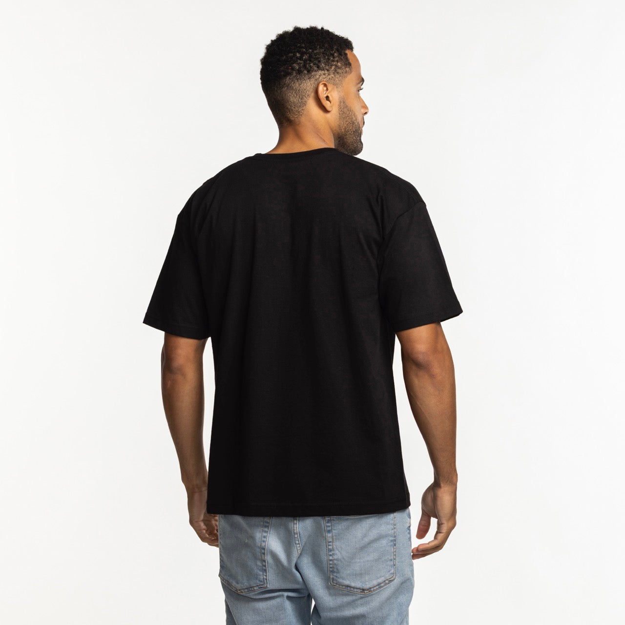 A man faces away from the camera, wearing a Mitchell & Ness Miami HEAT Mixed Up Tee by Mitchell & Ness and light blue jeans against a white background.