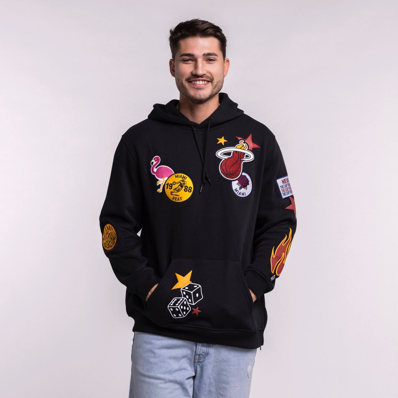 A smiling man with short dark hair wears the New Era Miami HEAT Up There Hoodie—a black standard fit hoodie by New Era with colorful patches—paired with light blue jeans against a plain light background.