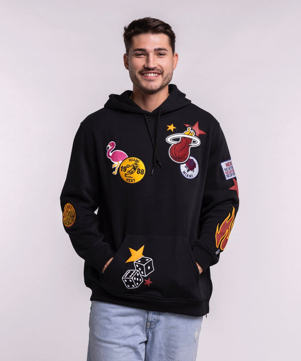 A smiling man with short dark hair wears the New Era Miami HEAT Up There Hoodie—a black standard fit hoodie by New Era with colorful patches—paired with light blue jeans against a plain light background.