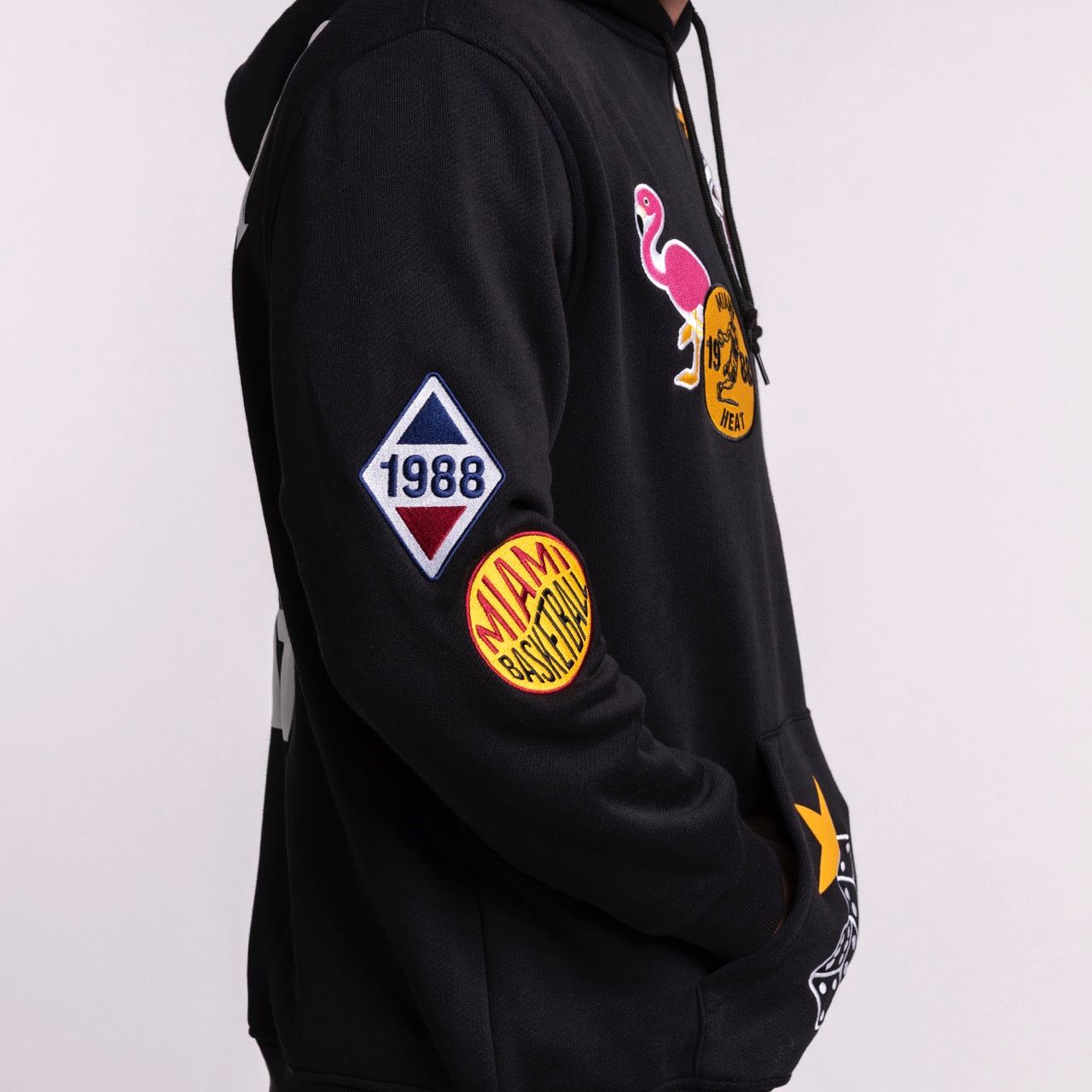 A person wears the New Era Miami HEAT Up There Hoodie—a black, standard-fit hoodie by New Era, featuring colorful patches like a 1988 diamond, yellow circle, and pink flamingo—made from polyester cotton and machine washable.