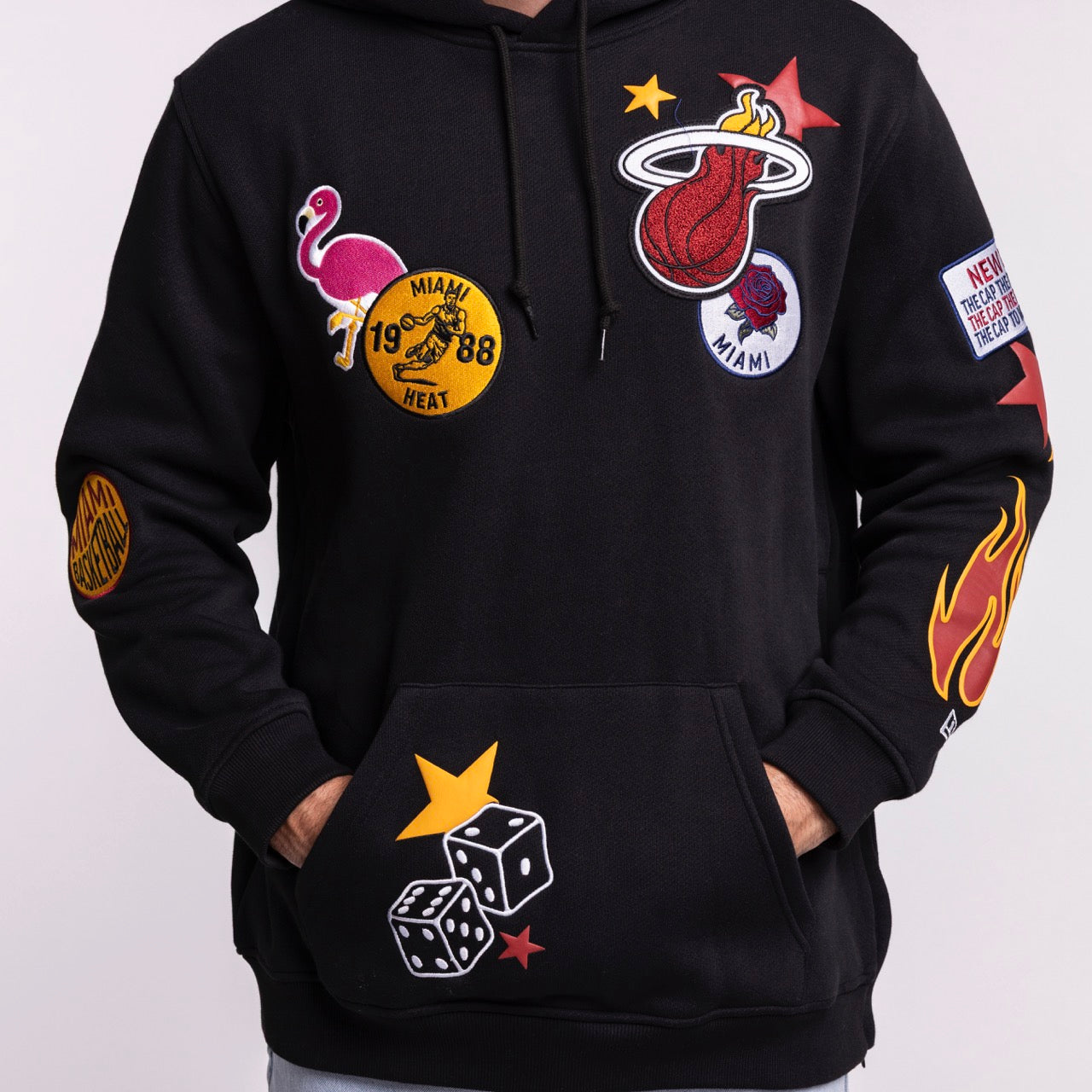 Wearing the New Era Miami HEAT Up There Hoodie, featuring colorful patches—flaming basketball, flamingo, star, dice, and Miami Heat logos. Standard fit, polyester-cotton blend, front pocket. Machine wash cold. By New Era.