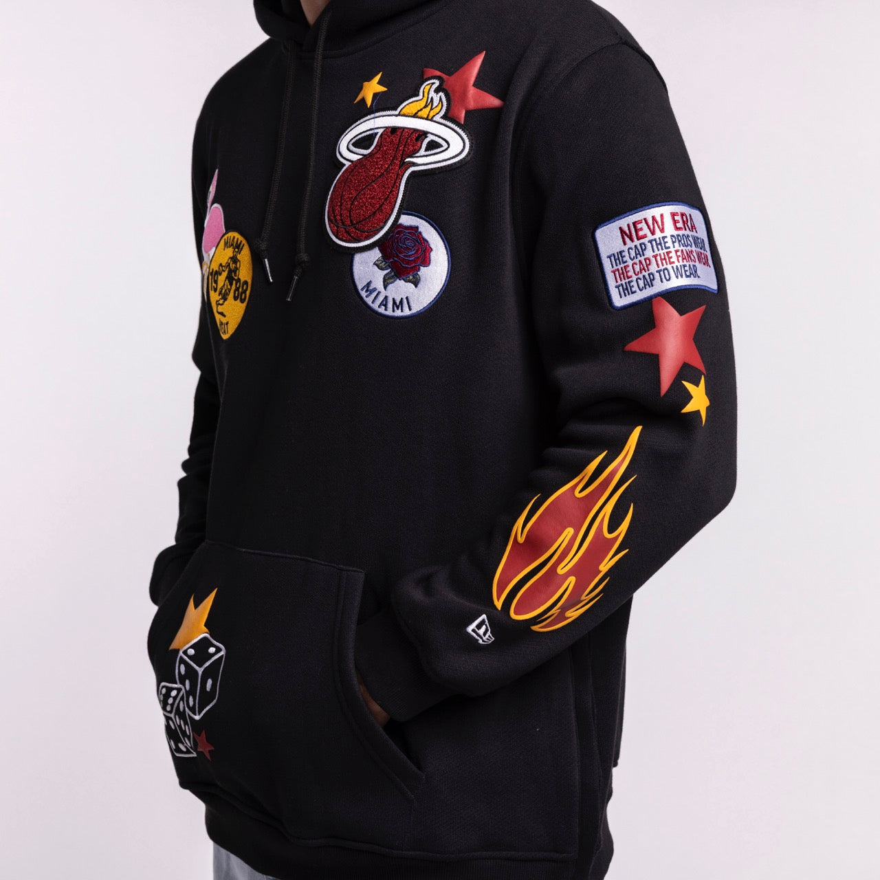A person wears the New Era Miami HEAT Up There Hoodie—a black, standard fit hoodie with Miami Heat logo, flaming basketball, rose, stars, dice, and “NEW ERA” patches. Polyester-cotton blend. Machine wash cold. Hands in hoodie pockets. Brand: New Era.