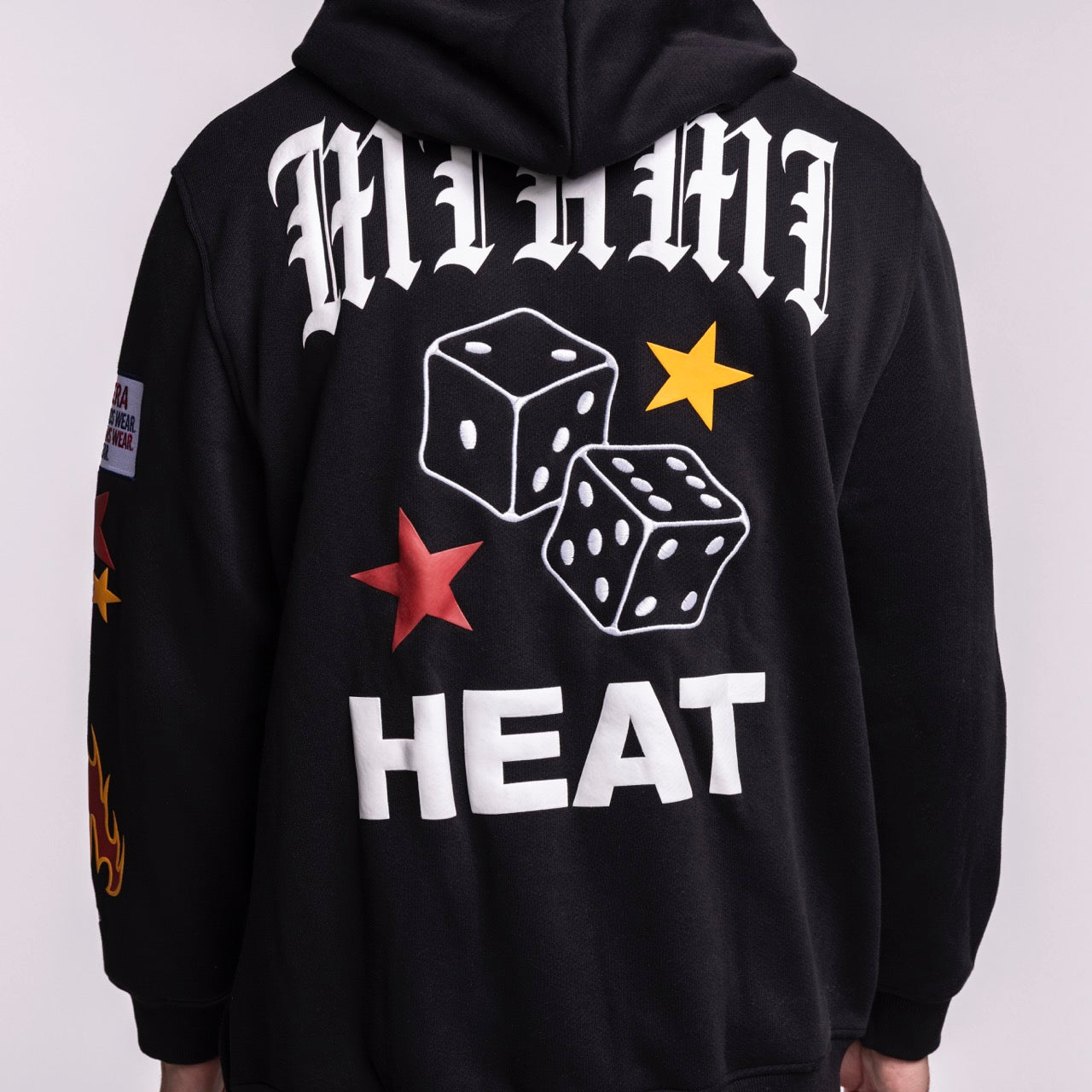 Wearing the New Era Miami HEAT Up There Hoodie, a standard fit black piece featuring bold white “MIAMI,” two dice, red and yellow stars, “HEAT” on the back, sleeve flames, and made from an easy-care polyester cotton blend.
