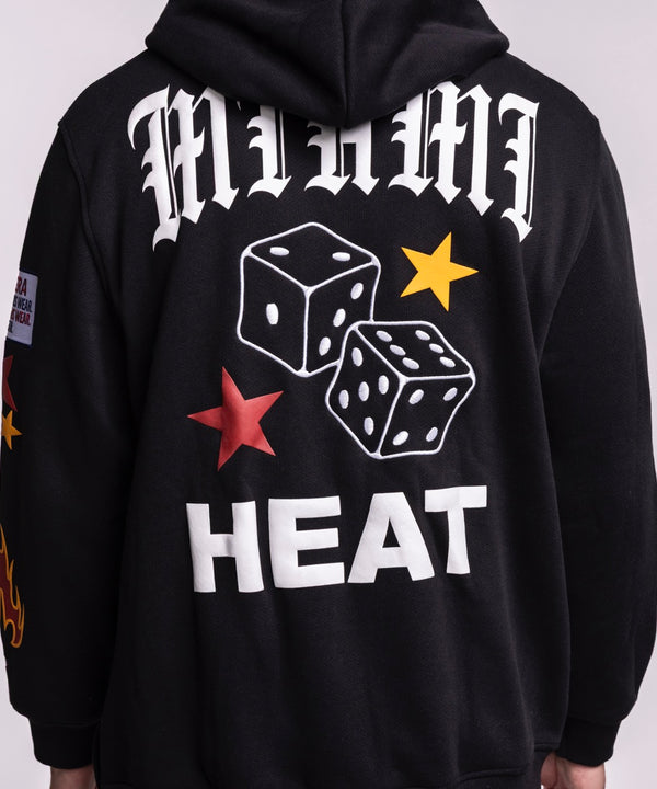 A smiling man with short dark hair wears the New Era Miami HEAT Up There Hoodie—a black standard fit hoodie by New Era with colorful patches—paired with light blue jeans against a plain light background.