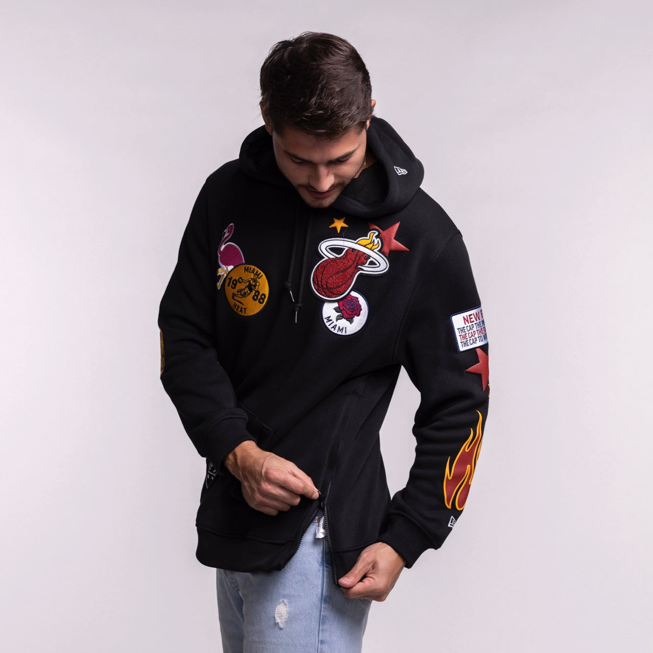 A man wearing the New Era Miami HEAT Up There Hoodie, featuring colorful patches like a basketball team logo, stars, and a flame, adjusts his sleeve. This soft polyester-cotton hoodie by New Era is styled with light blue ripped jeans.