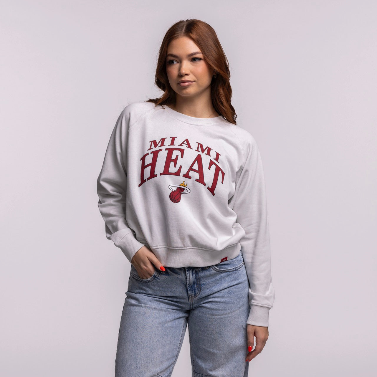 A woman with long brown hair wears the Sportiqe Miami HEAT Wordmark Women's Crewneck by Sportiqe and blue jeans, standing with one hand in her pocket against a plain light background.
