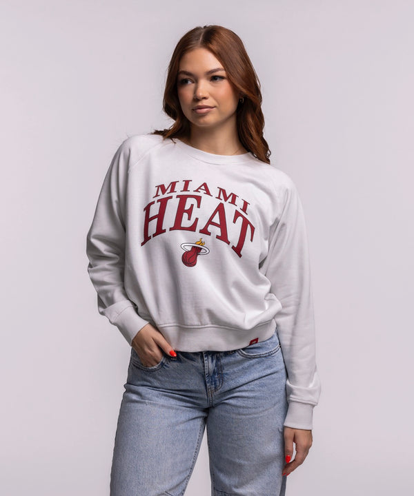 A woman with long brown hair wears the Sportiqe Miami HEAT Wordmark Women's Crewneck by Sportiqe and blue jeans, standing with one hand in her pocket against a plain light background.