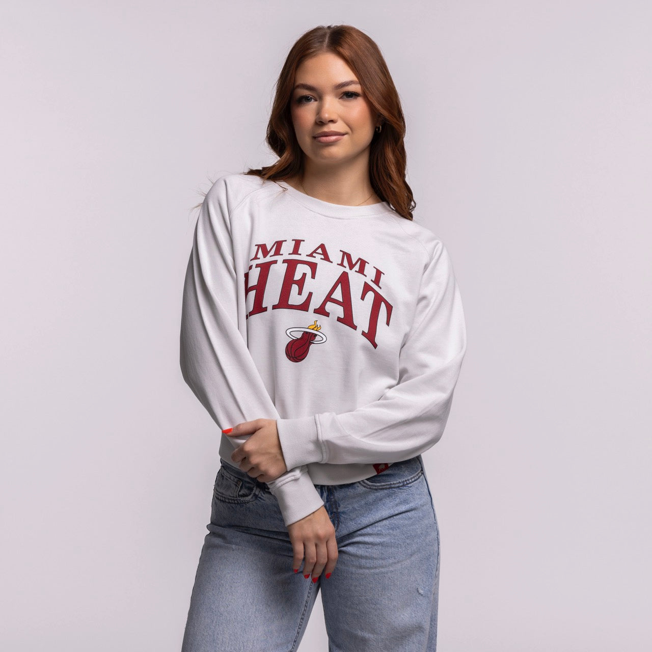 A woman with long brown hair wears the Sportiqe Miami HEAT Wordmark Women's Crewneck in a relaxed cotton fit and light blue jeans, standing against a light gray background with a relaxed expression. Machine wash cold for best care. Brand: Sportiqe.