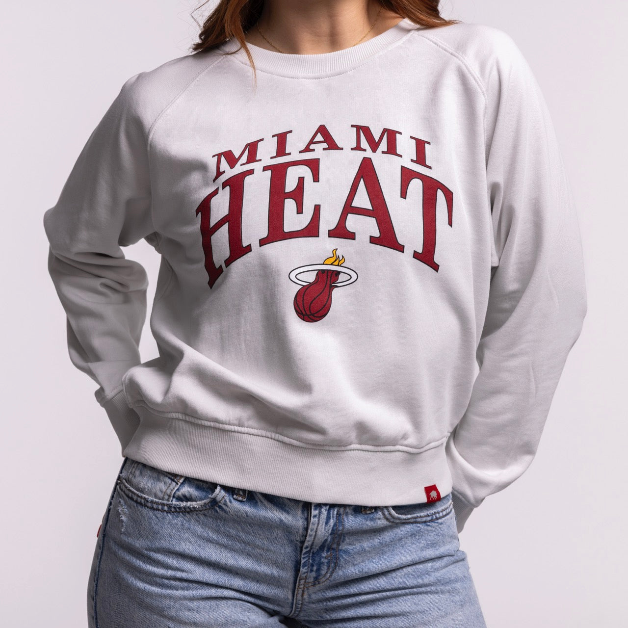 A woman wears the Sportiqe Miami HEAT Wordmark Women's Crewneck by Sportiqe—a white, cotton, relaxed-fit sweatshirt with MIAMI HEAT and the flaming logo on the front—paired with light blue jeans; it's machine washable.