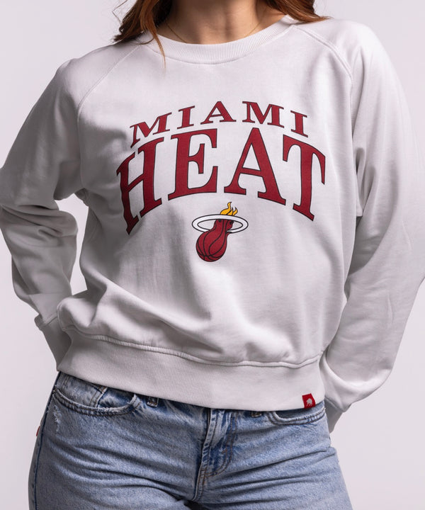 A woman with long brown hair wears the Sportiqe Miami HEAT Wordmark Women's Crewneck by Sportiqe and blue jeans, standing with one hand in her pocket against a plain light background.