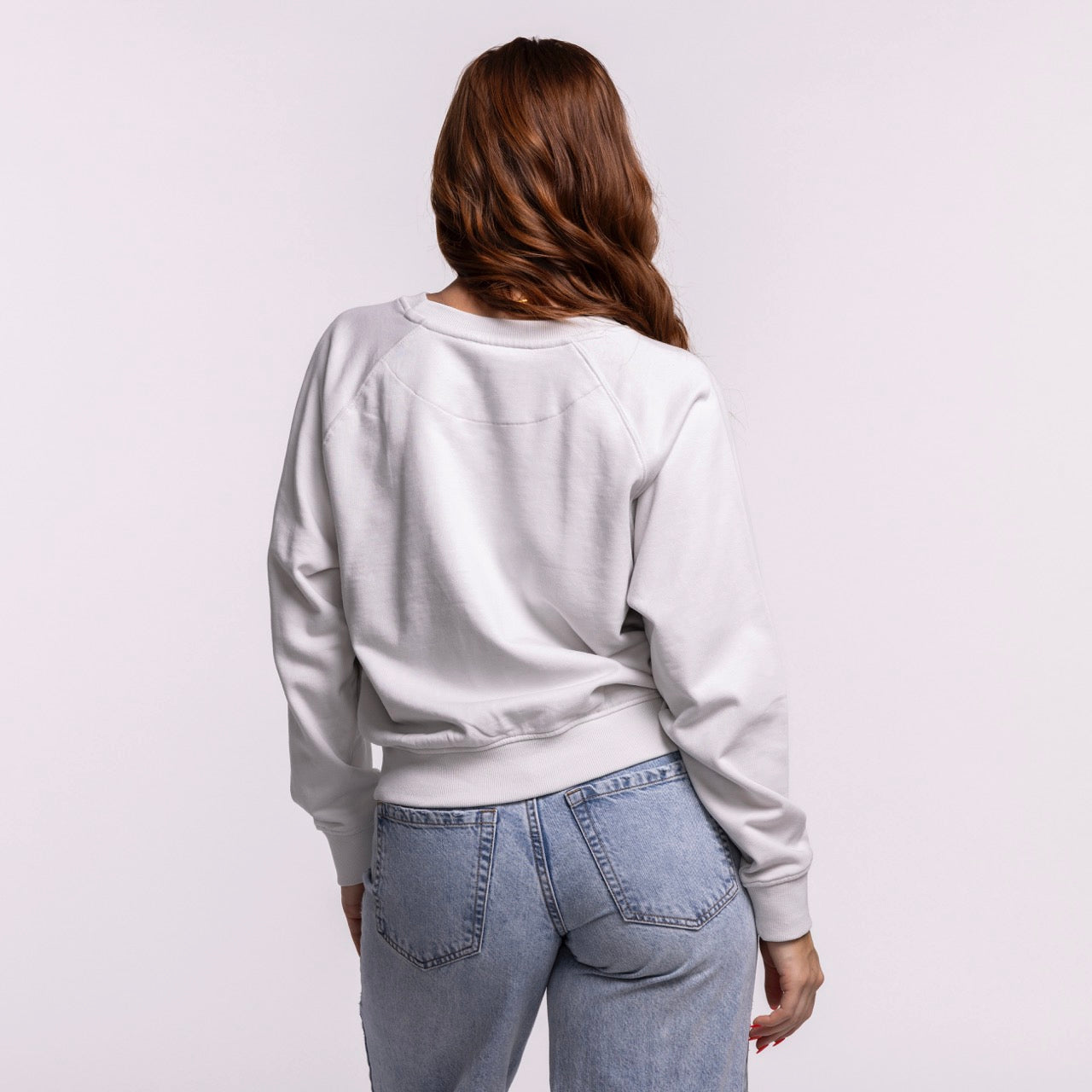 A woman with long, wavy brown hair stands with her back to the camera, wearing the Sportiqe Miami HEAT Wordmark Women's Crewneck by Sportiqe and high-waisted blue jeans against a plain light background.