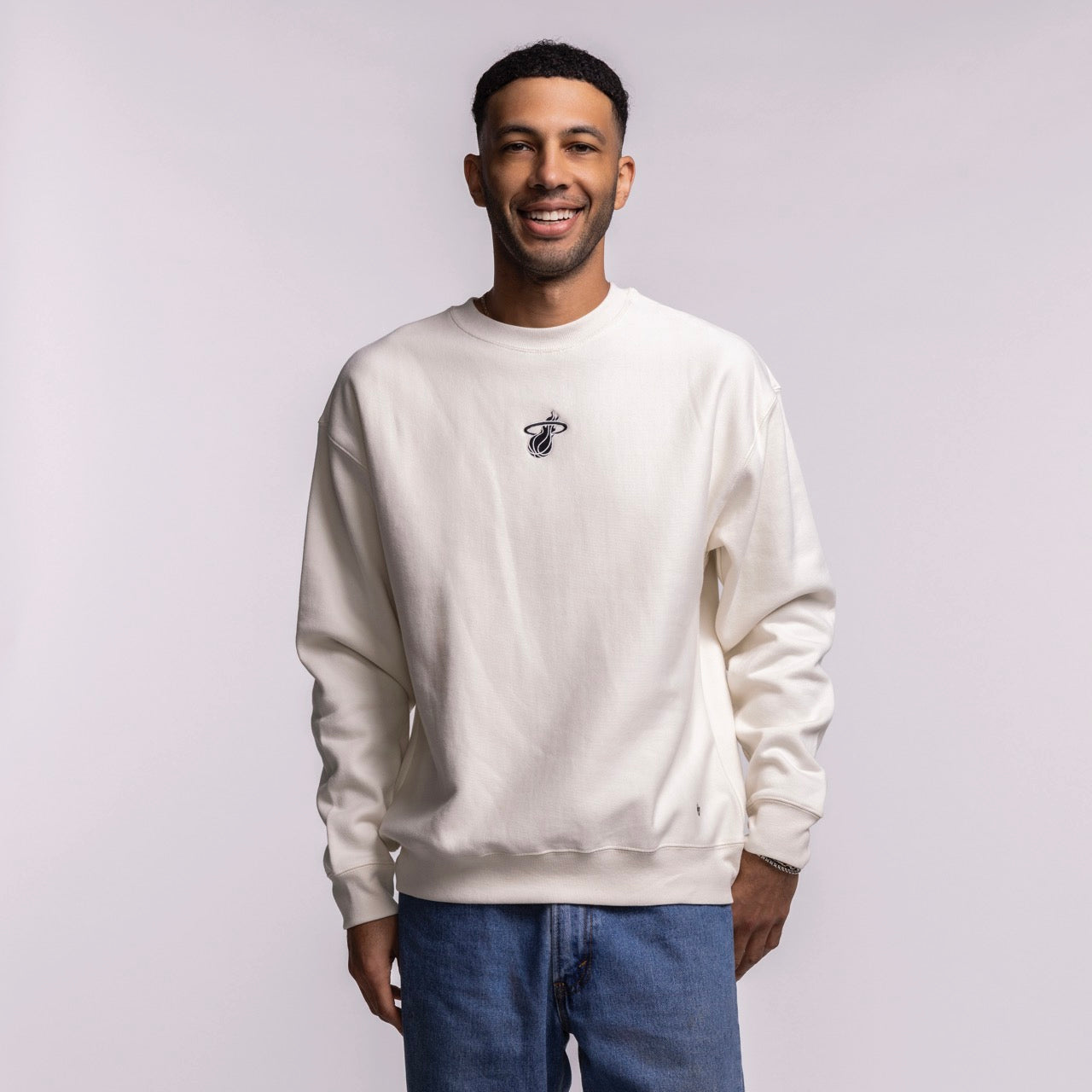 A man smiles against a light background, wearing the '47 Brand Miami HEAT Cream Logo Foundation Crewneck with a soft brushed interior and small embroidered logo on the chest, paired with blue jeans.