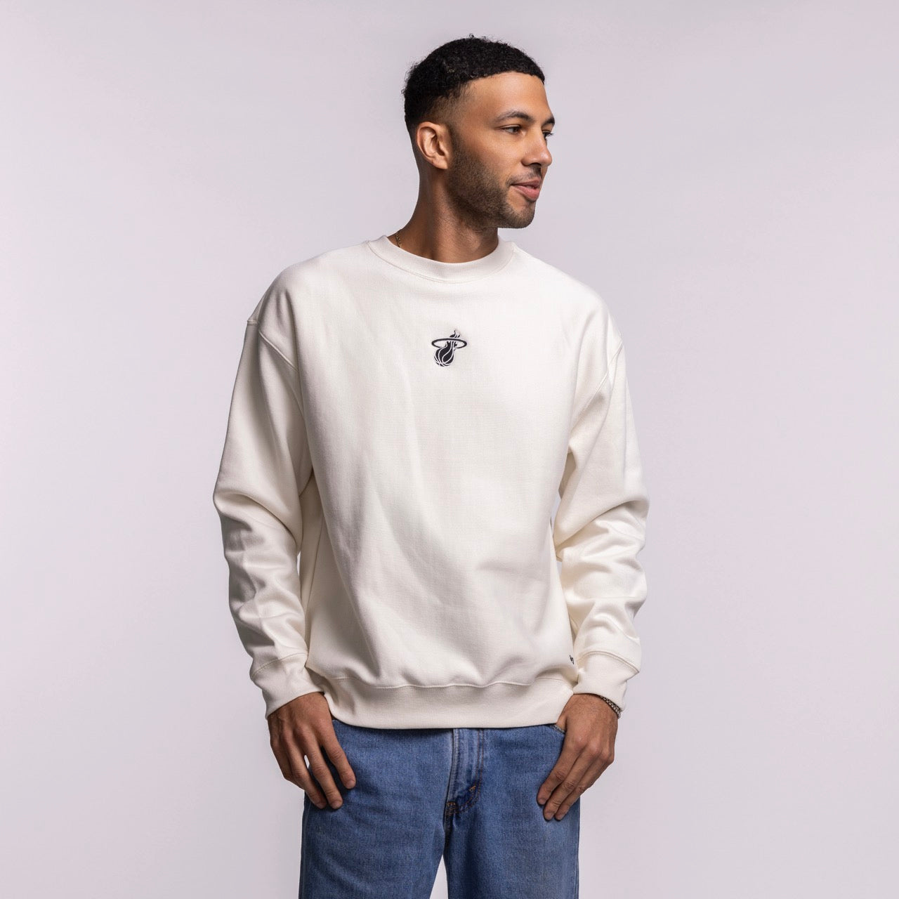 A man wearing a '47 Brand Miami HEAT Cream Logo Foundation Crewneck and blue jeans stands against a light gray background, looking slightly to his left with a relaxed expression.