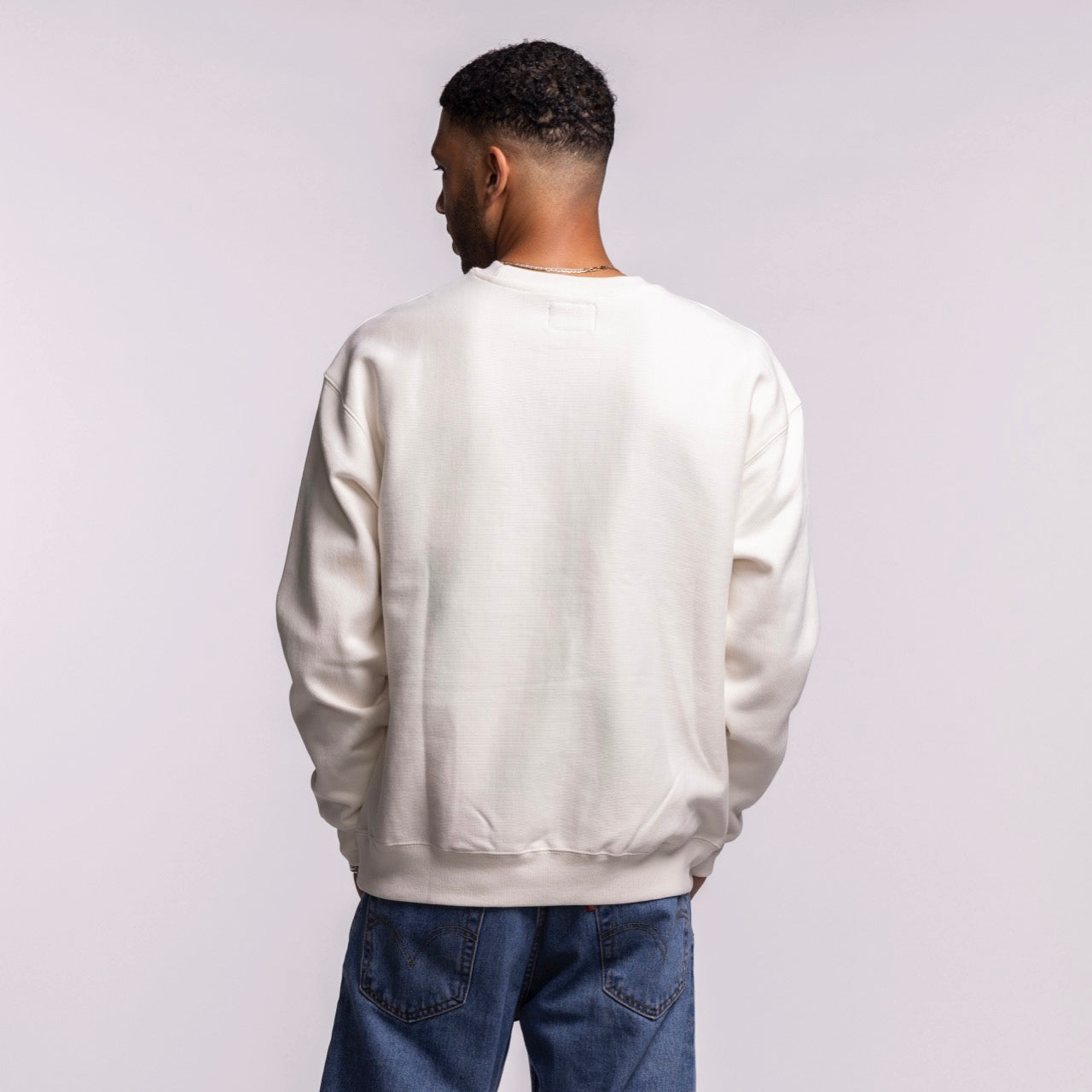 A man faces away from the camera, wearing the '47 Brand Miami HEAT Cream Logo Foundation Crewneck with a soft brushed interior and blue jeans, set against a light gray background.