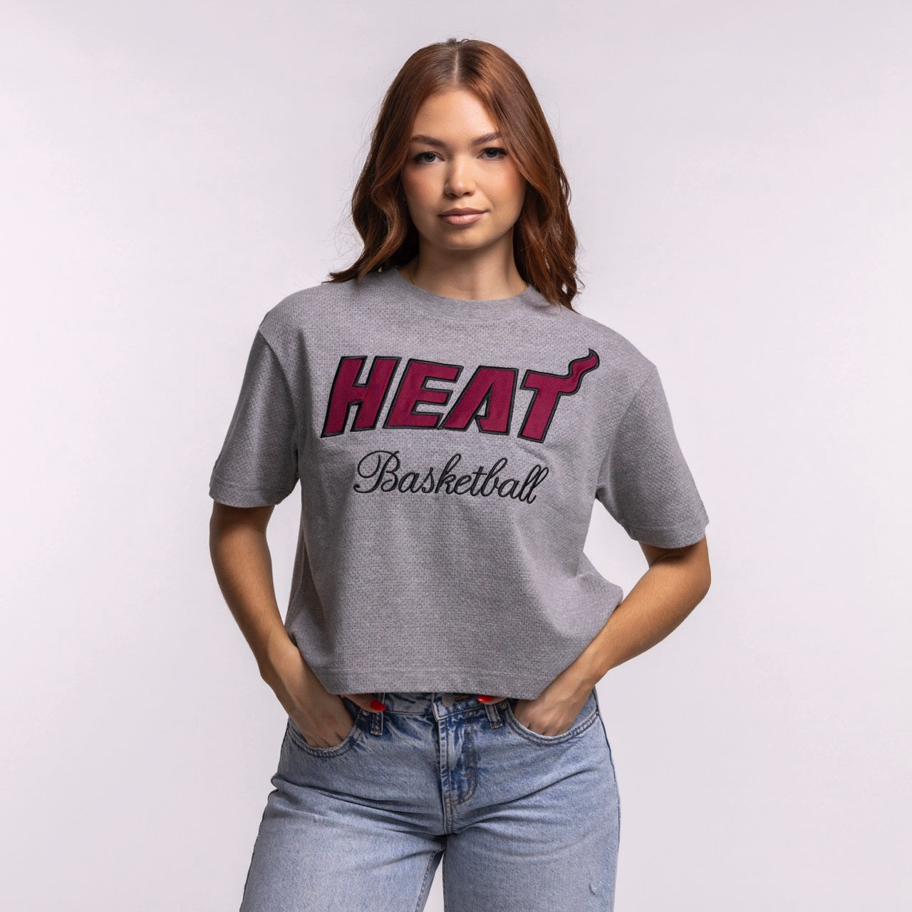 A woman with long brown hair wears the '47 Brand Miami HEAT Pivot Women's Cropped Tee, a relaxed gray cotton-polyester t-shirt with red and black 